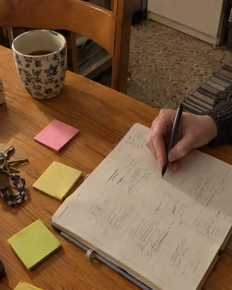 Mani che scrivono su un'agenda settimanale, con post-it e un timer da cucina sul tavolo